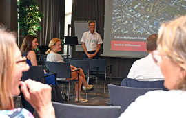 Teilnehmerinnen und Teilnehmer eines Diskussionsforums sitzenund stehen vor einer Projektionsleinwand