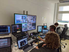 Mehrere Menschen sitzen in einem Technikraum vor Laptops und nehmen an einer Videokonferenz teil.
