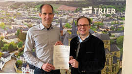 Thilo Becker und Ralf Britten stehen vor einer Luftaufnahme der Trierer Innenstadt und präsentieren ein offizielles Schreiben