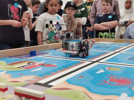 Kinder stehen rund um einen Wettbewerbstisch und schauen einem Roboter zu