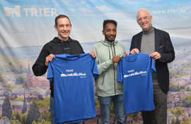 Wolfram Leibe überreicht Samuel Fitwi und dessen Trainer Yannick Duppich als Willkommensgruß blaue Laufshirts mit Trier-Motiven.