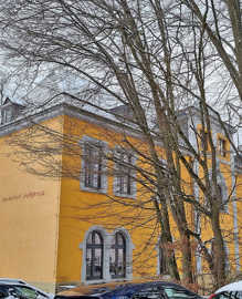 Auf einem großen Gebäude mit gelber Fassade ist links an der Hauswand der Schriftzug "Kinderhort Heiligkreuz" zu lesen. Im Vordergrund stehen einige hohe Bäume.