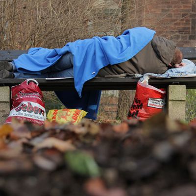 Eine Person liegt zugedeckt auf einer Parkbank, vor ihr auf dem Boden volle Plastiktüten